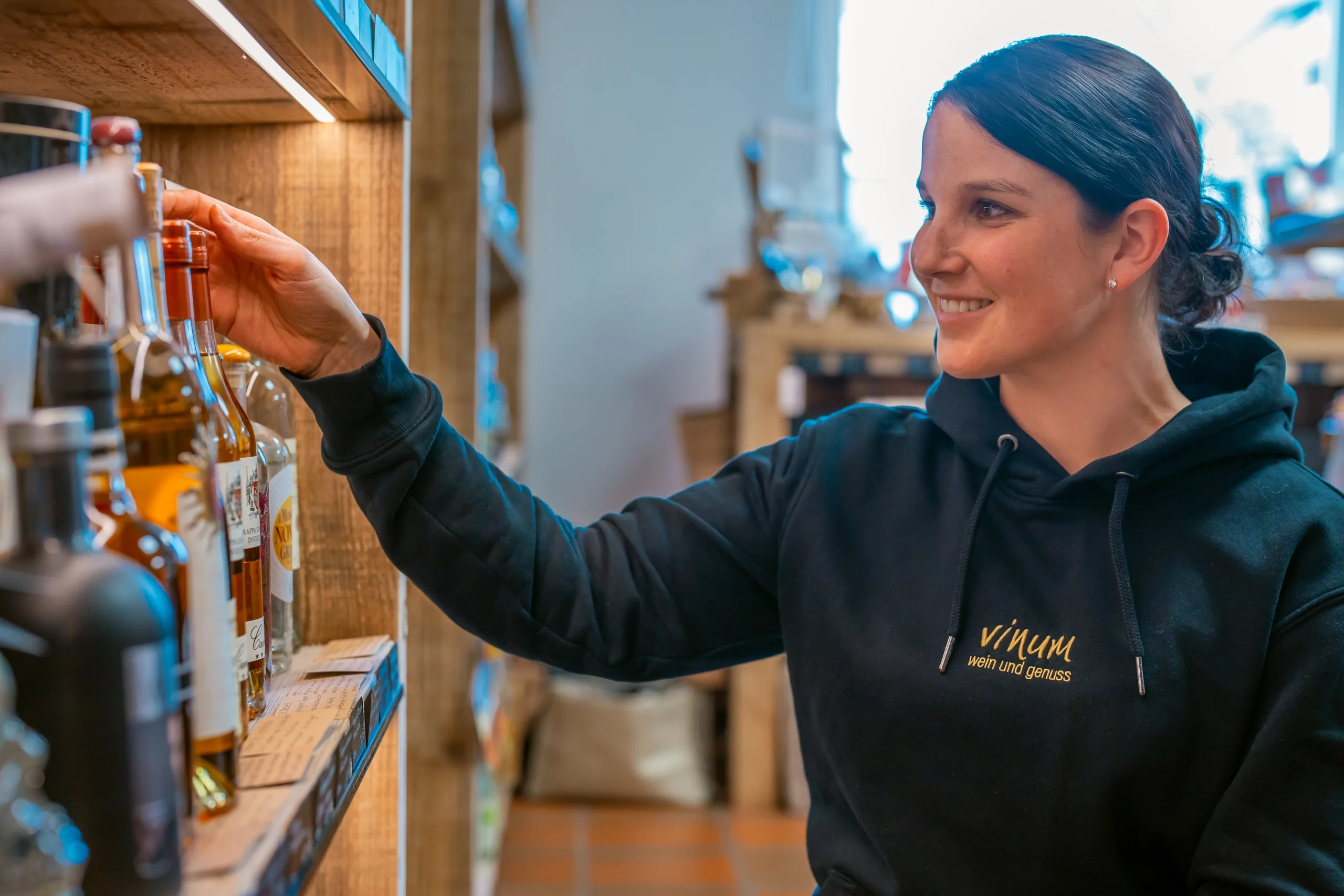Eine Frau mit einem schwarzen Kapuzenpullover mit der Aufschrift „Vinum“ lächelt und greift nach einer Flasche auf einem Holzregal in einem Geschäft. Um sie herum sind Flaschen in den Regalen aufgereiht. Der Laden ist innen sanft beleuchtet.