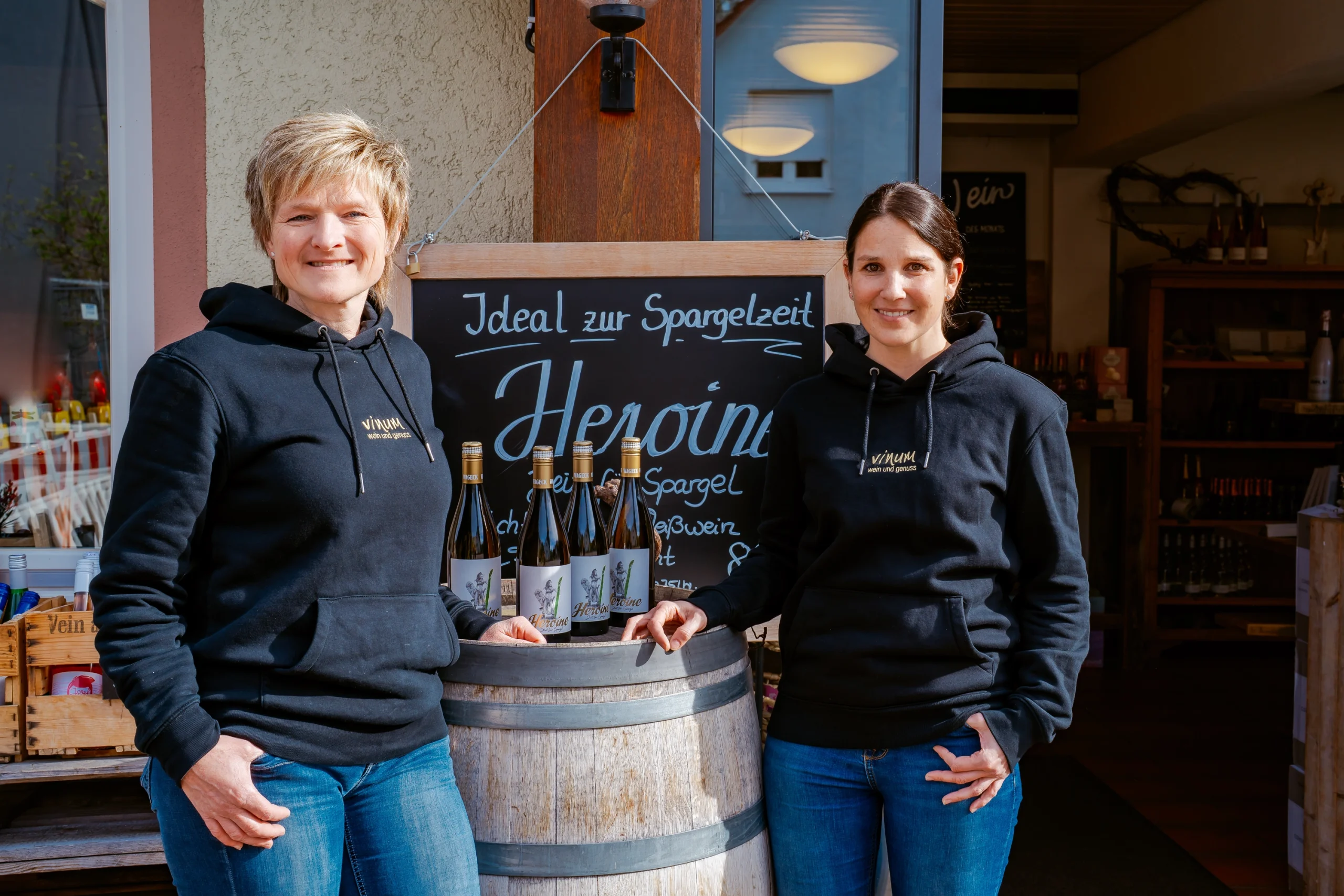 Zwei Frauen in schwarzen Kapuzenpullovern stehen neben einem Holzfass, in dem Weinflaschen ausgestellt sind, vor einem Geschäft. Auf einer Tafel hinter ihnen steht „Ideal zur Spargelzeit und Heppni“. Die Atmosphäre ist leger und einladend.