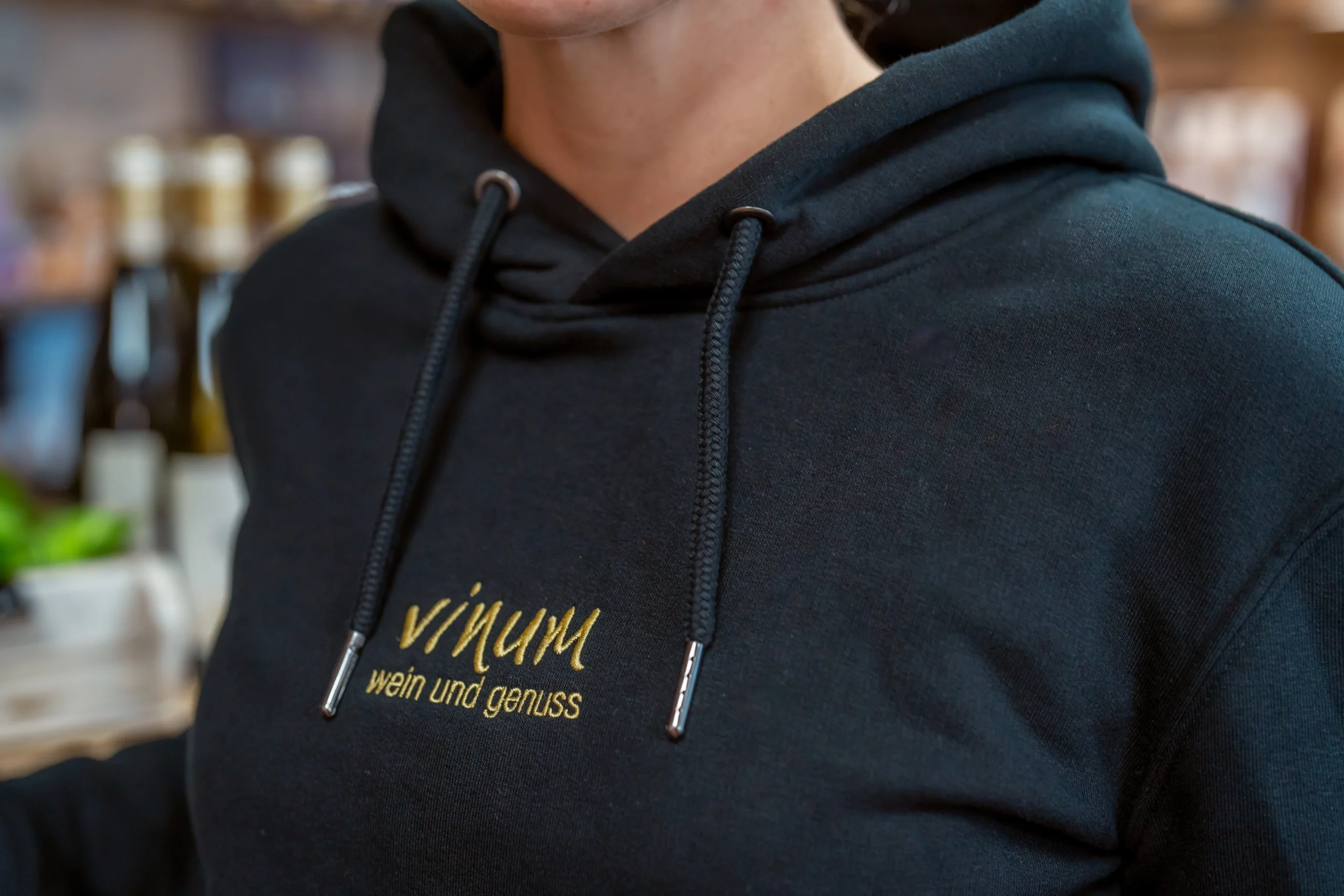 Nahaufnahme einer Person, die einen dunklen Kapuzenpullover mit gelbem Text trägt, auf dem „vinum wein und genuss“ steht. Weinflaschen sind im Hintergrund verschwommen.