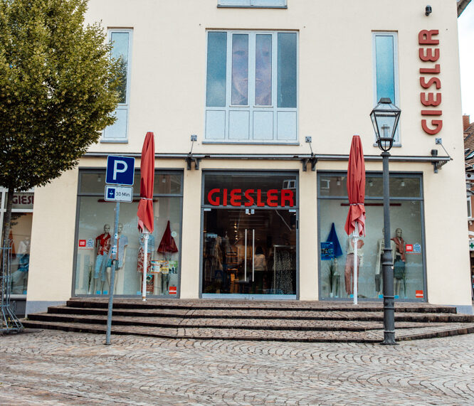 Ein creme farbenes Gebäude mit rotem Spitzdach und der Aufschrift GIESLER mit gekleideten Puppen im Schaufenster und zwei roten Schirmen vor dem Eingang.
