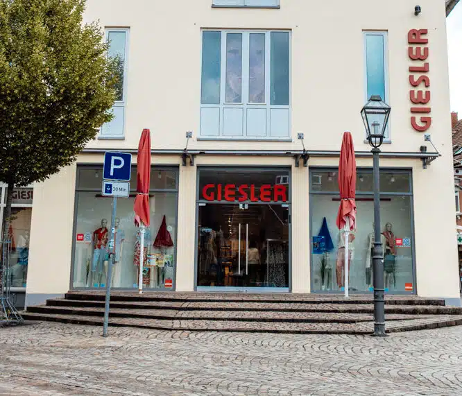 Ein creme farbenes Gebäude mit rotem Spitzdach und der Aufschrift GIESLER mit gekleideten Puppen im Schaufenster und zwei roten Schirmen vor dem Eingang.