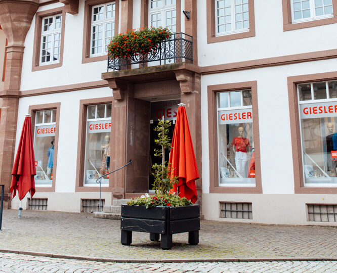 Ein weißes Gebäude mit braun umrandeten Fenstern und Spitzdach mit der Aufschrift GIESLER mit gekleideten Puppen im Schaufenster und zwei roten Schirmen vor dem Eingang.