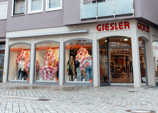 Ein graues Gebäude mit Spitzdach und der Aufschrift GIESLER trend sowie gekleideten Puppen im Schaufenster.
