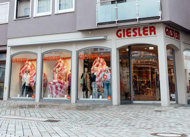 Ein graues Gebäude mit Spitzdach und der Aufschrift GIESLER trend sowie gekleideten Puppen im Schaufenster.