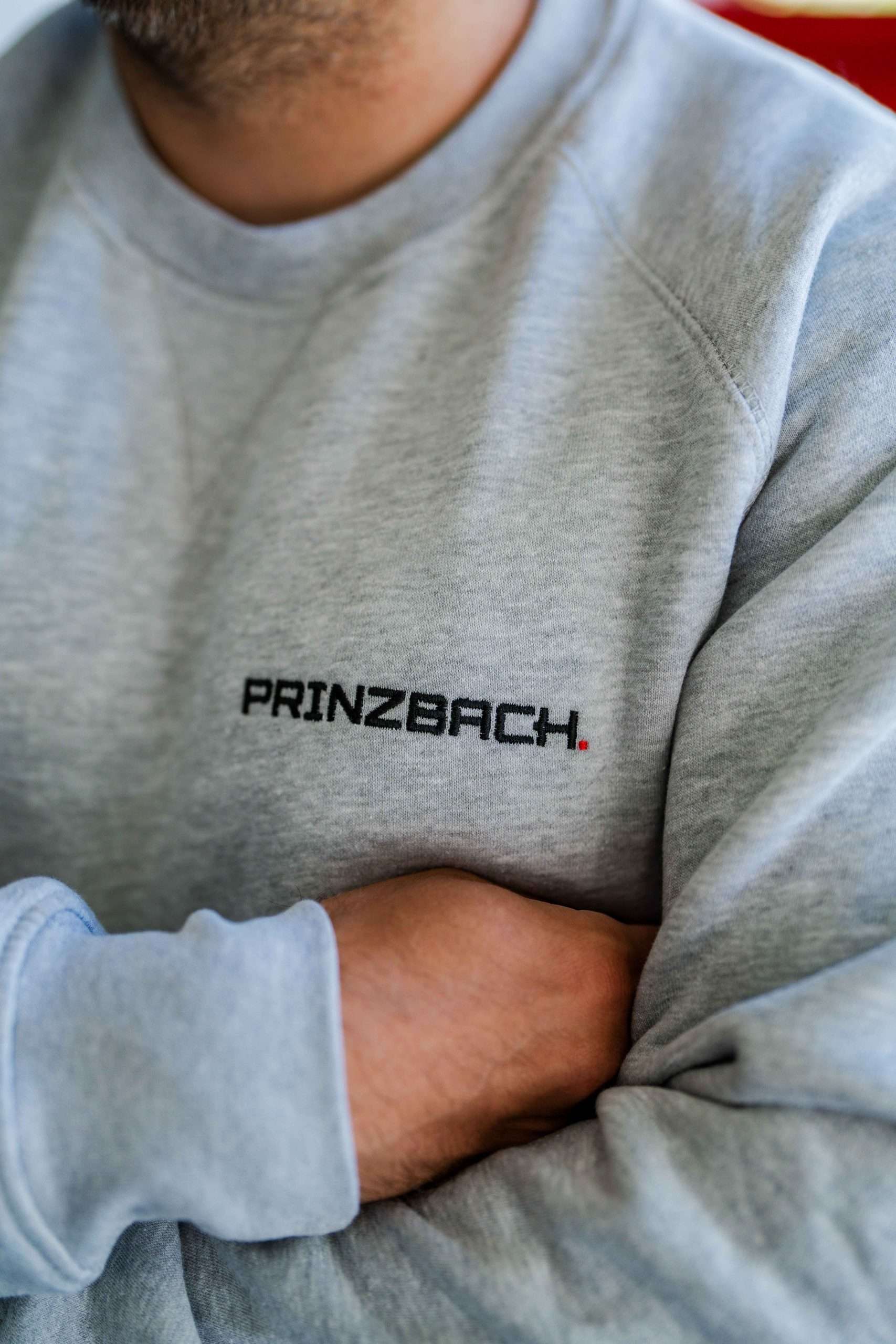 Nahaufnahme einer Person, die ein hellgrauen Pullover mit dem schwarz aufgestickten Wort PRINZBACH trägt. Der Mann im Pullover steht vor einem unscharfen Hintergrund.