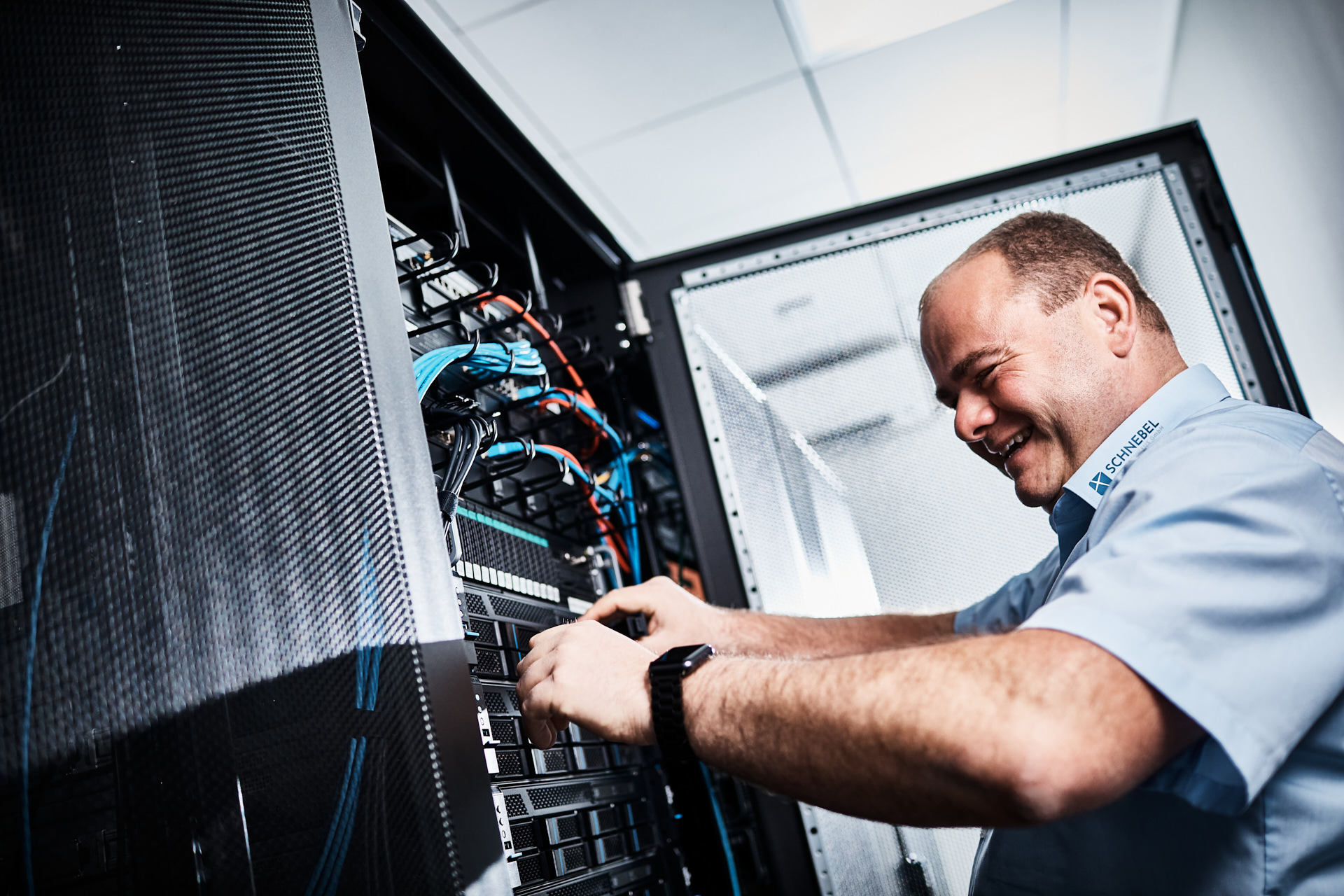 Ein Mann im hellblauen Hemd arbeitet an einem Server. Dabei lacht er und trägt eine Smartwatch.