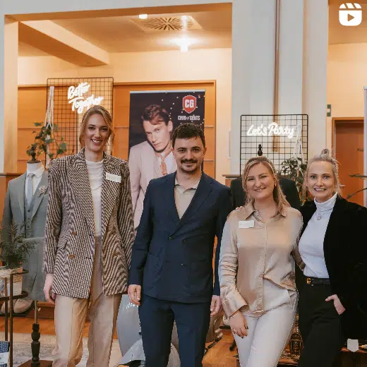 4 Personen stehen in einem Kaufhaus und lächeln. Davon sind es 3 Frauen und 1 Mann. Die linke Frau trägt einen Blazer in den Farben beige und schwarz. Der Mann einen dunkelblauen Anzug, die Frau rechts daneben ein beiges Hemd und die Frau ganz rechts einen schwarzen Anzug.