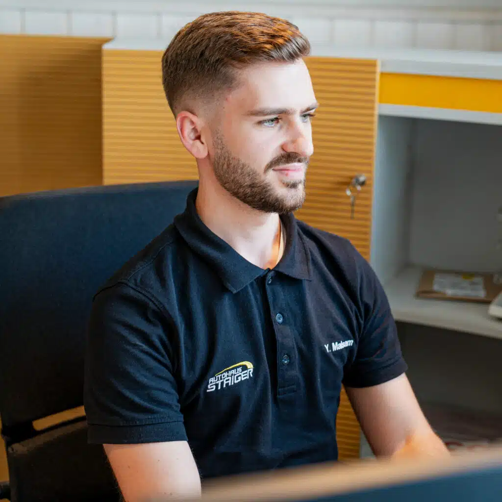 Ein junger Mann sitzt in einem Bürogebäude auf einem schwarzen Bürostuhl mit einem schwarzen Polo und der Aufschrift Autohaus Staiger