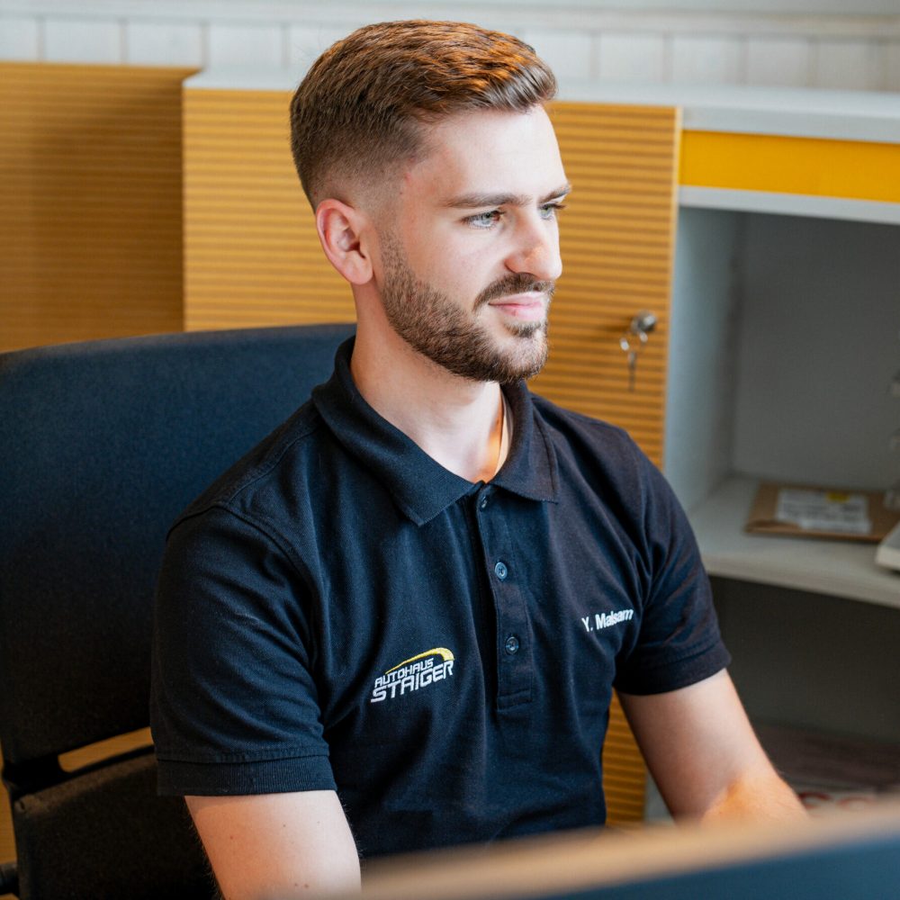 Ein junger Mann sitzt in einem Bürogebäude auf einem schwarzen Bürostuhl mit einem schwarzen Polo und der Aufschrift Autohaus Staiger